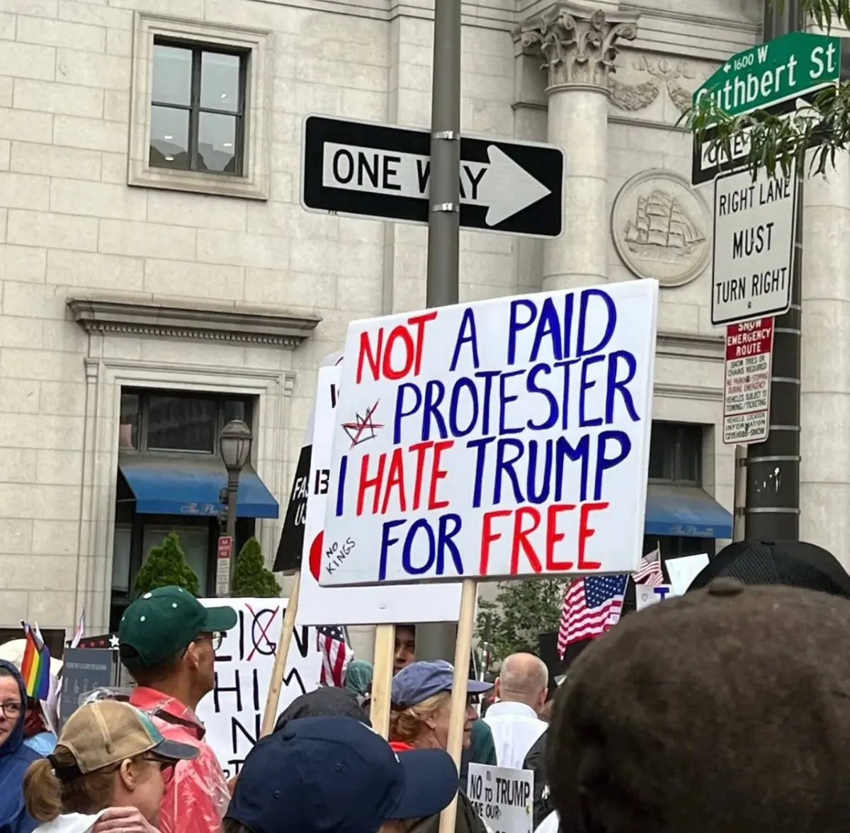 Street protest with a large anti-Trump sign reading 'NOT A PAID PROTESTER I HATE TRUMP FOR FREE' in an urban setting
