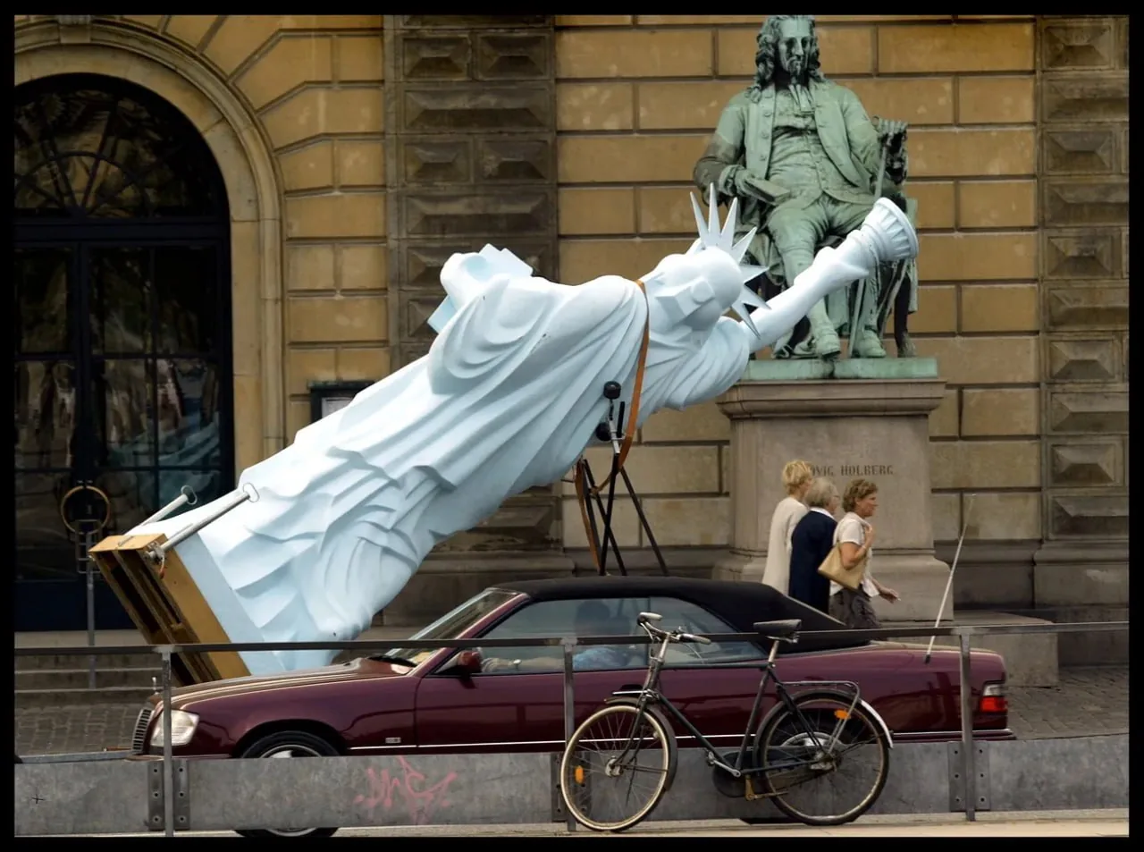 Street scene with a toppled Statue of Liberty sculpture, pedestrians, a parked car, and a bicycle by a building