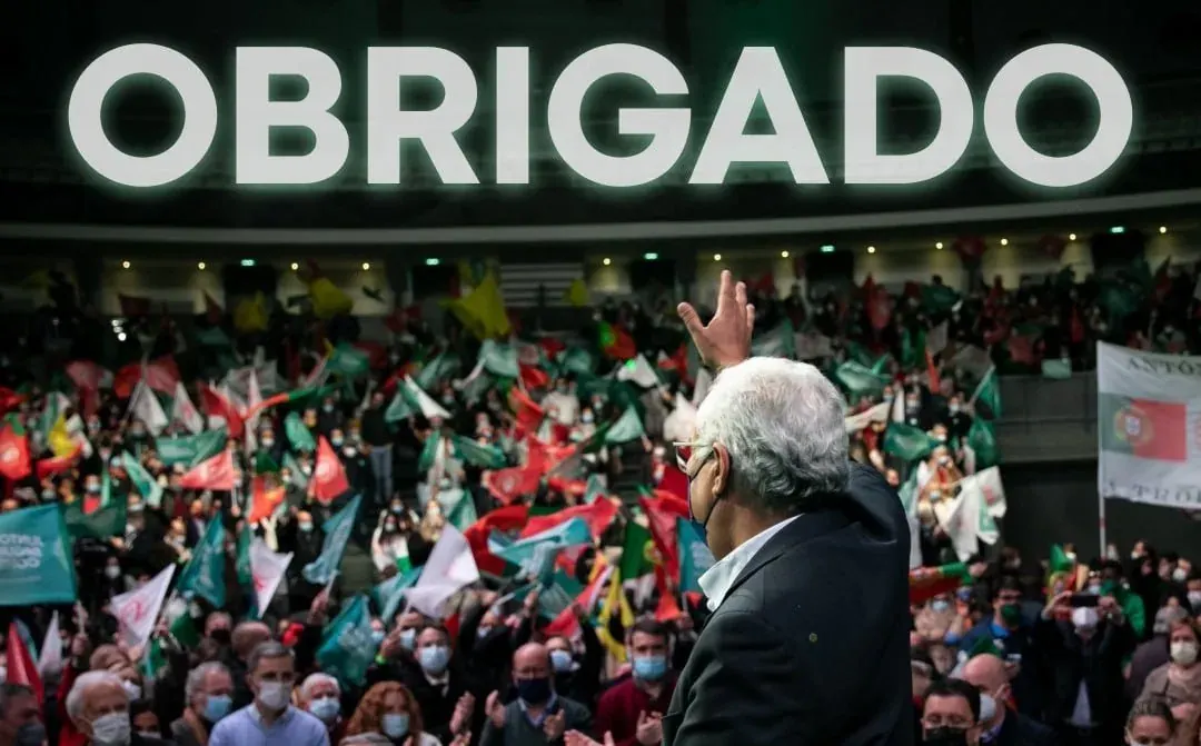 Public speaker addressing a large masked crowd with flags and a banner reading OBRIGADO