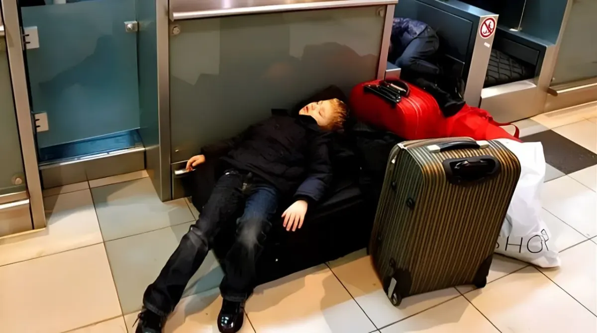 Tired traveler lying on the floor beside luggage in an airport check-in area during a long transit